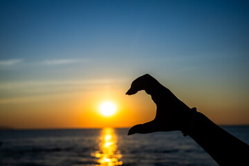 silhouetted hand forming a half-heart gesture in front of a glowing sunset over the ocean © pandaclub23