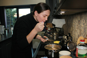 A woman at home, going about her daily business. A woman is making soup in the kitchen, a woman is salting and tasting the broth.