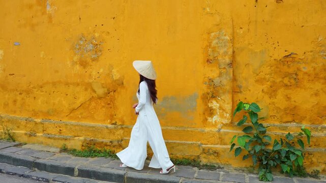 4k slow motion video of Asian woman wearing white Vietnamese cultural dress and traditional conical hat walking around Hoi An, Vietnam, with yellow walls as a backdrop.
