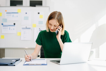Business woman talking on the phone while writing notes and working on a laptop in a bright modern office.