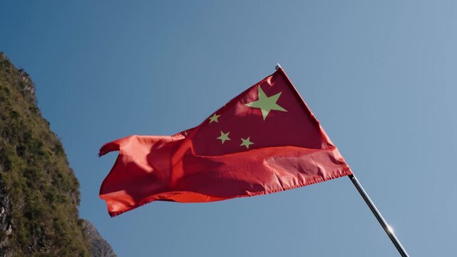The Chinese flag ripples in bright daylight as steep karst cliffs rise beneath clear sky.