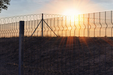Dramatic golden hour sunlight silhouettes a high-security fence topped with razor wire, evoking...