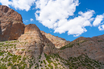 Upward view of massive, layered, reddish-brown rock cliffs and slopes with patches of green shrubs, set against a bright blue sky with white clouds.