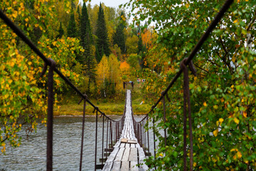 A narrow wooden suspension bridge stretching across a flowing river toward a dense forest featuring vibrant yellow and green autumn foliage
