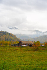 An old, rustic wooden house stands in a grassy meadow with rolling mountains in the background covered in autumn foliage and obscured by low-hanging clouds