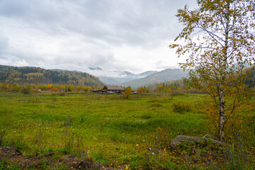 A traditional, long wooden house or log cabin with a pitched roof sits in a vast, green meadow surrounded by hills covered in yellow and green autumn forest