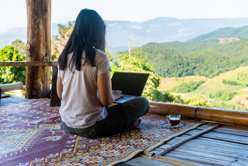 Asian woman digital nomad working on laptop outdoors on a wooden balcony with stunning mountain view, Concept of remote work, freedom, and work-life balance.