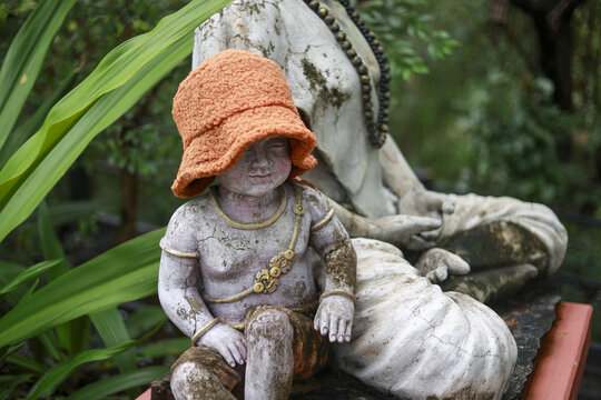 Peaceful stone statue of child wearing bright orange hat, weathered art decoration sitting quietly in lush green garden. pensive figure evokes sense of tranquility - Powered by Adobe