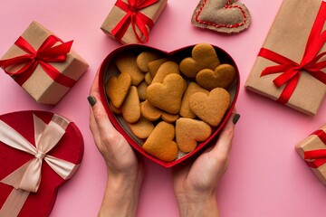 Hands holding a heart shaped box overflowing with delicious heart shaped cookies surrounded by gifts