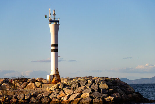 Prominent Navigation Lighthouse with White and Black Stripes, Standing on a Detailed Rocky Breakwater Under Clear Daylight, Overlooking the Open Sea.
