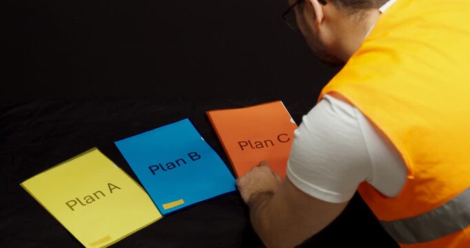 A worker in a safety vest stands over three folders labeled Plan A, Plan B, and Plan C, thinking carefully and appearing uncertain which plan to choose.