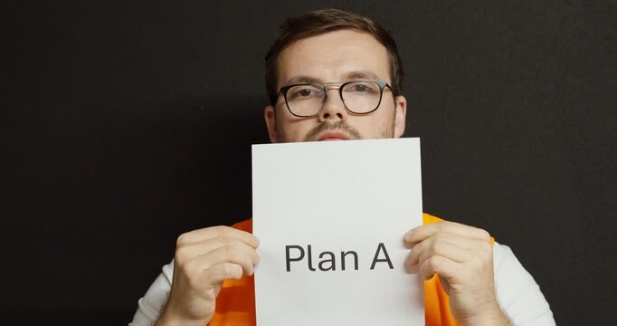 A man in an orange safety vest holds up a paper labeled &ldquo;Plan A&rdquo; in front of a black background, representing decision-making and planning at work.