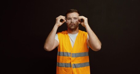 A worker in a high-visibility vest holds and puts on safety goggles, demonstrating proper workplace safety and protection practices.