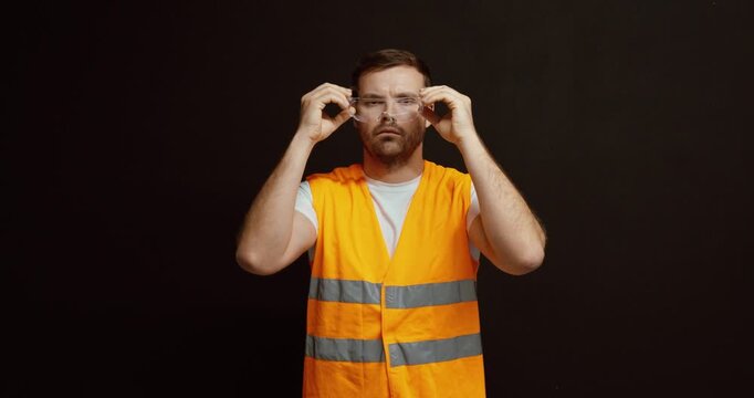 A worker in a high-visibility vest holds and puts on safety goggles, demonstrating proper workplace safety and protection practices.