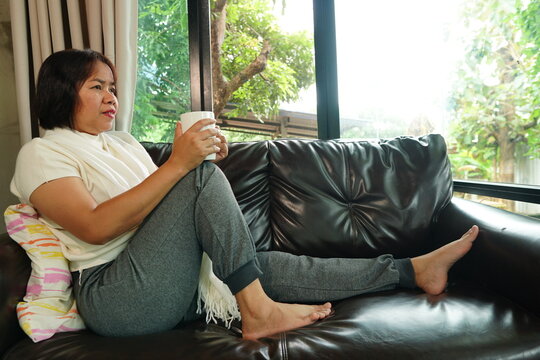 A woman is sitting and relaxing on a brown leather sofa by a large window, holding a coffee mug and looking outside.