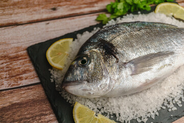 Whole raw sea bream fish presented on a black slate, surrounded by coarse salt crystals and fresh lemon slices