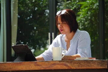 A woman is sitting, working with a tablet and a coffee mug next to her, by a window with natural light.
