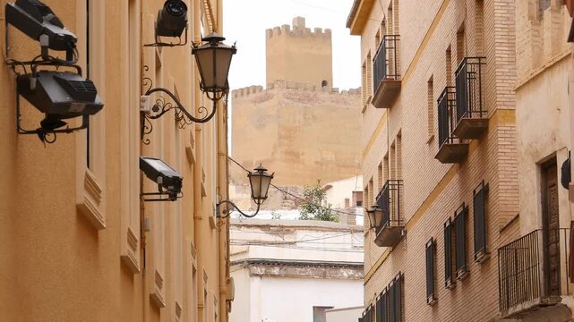 The tower of the Alcazaba of Guadix is ​​framed between two old buildings on a narrow street. The traditional ochre architecture and period lampposts contrast with modern spotlights.