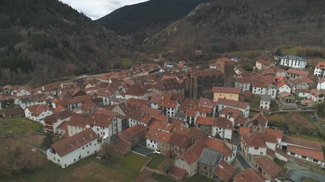 Aerial ascending orbit around Isaba village featuring tall central tower and old buildings surrounding on a cloudy day.
