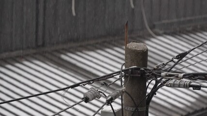 Rain falls over a worn concrete utility pole with tangled electrical cables and connectors, while a corrugated metal roof in the background forms soft lines and muted tones in the dim overcast weather - Powered by Adobe
