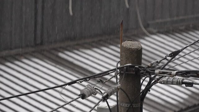Rain falls over a worn concrete utility pole with tangled electrical cables and connectors, while a corrugated metal roof in the background forms soft lines and muted tones in the dim overcast weather