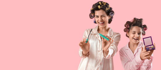 Little girl and her mother with hair curlers in pajamas doing makeup on pink background