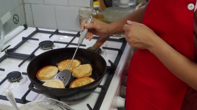 A cook in a red apron flips golden pancakes with a metal spatula. She fries the batter in a hot black skillet on a white gas stove in a home kitchen.