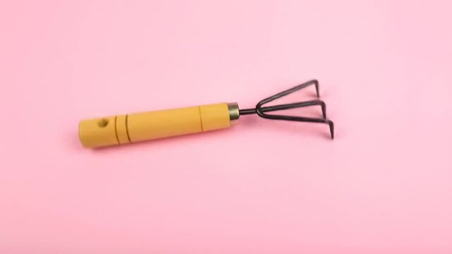 A small gardening tool with a wooden handle rests on a pink surface. Its tines are facing left