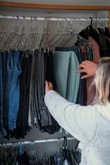 A person reaches into an organized closet, selecting a pair of pant hanging on a wire hanger under soft indoor light.