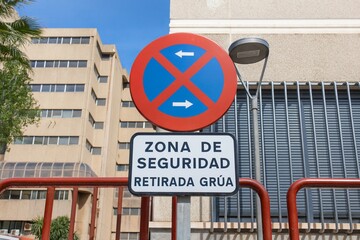 High-resolution photograph of a Spanish no-parking and tow-away safety zone sign against an urban backdrop, featuring modern buildings
