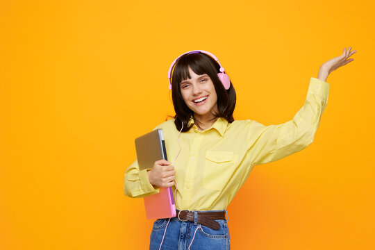 Cheerful student wearing a yellow shirt and pink headphones dances against a bright orange background, holding a notebook and smiling in a lively creative moment.