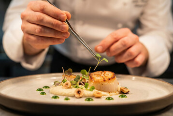 Chef using tweezers to garnish seared scallop dish with microgreens in fine dining restaurant