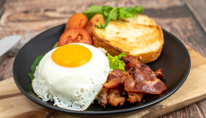 Sunny side up egg bacon toast tomato lettuce breakfast meal on black plate wooden table