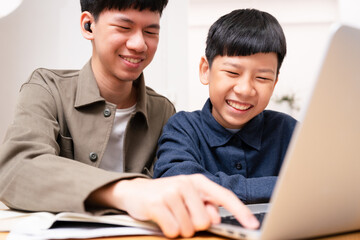 Two Asian sibling study online learning, teenage brother teach younger boy with laugh and smile at home, looking at computer laptop, having fun, happiness and joy. Education and e-learning technology