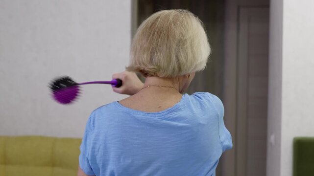 Senior woman wearing a blue t shirt is massaging her back and shoulders with a special purple tool, performing various movements to relieve muscle tension and improve blood circulation