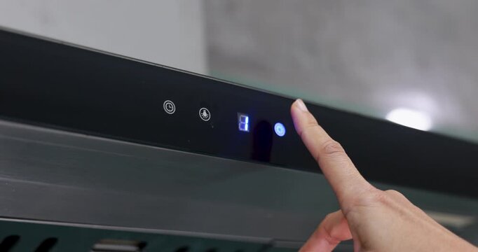 A hand presses a glowing blue touch kitchen hood control button on the digital display of a modern black kitchen range hood, adjusting the fan speed for cooking ventilation. 4k VDO.