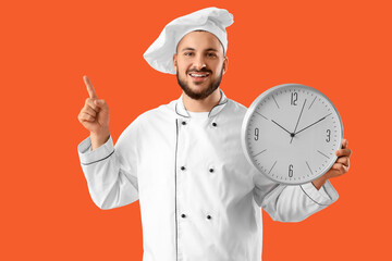 Young happy male chef with big clock pointing at something on orange background
