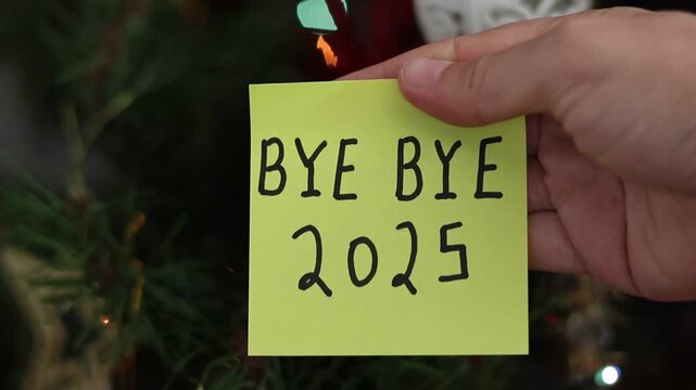 Un primer plano de la mano de una mujer con una nota de color amarillo  con la palabra adi&oacute;s 2025 en un fondo de luces de colores. Fin de a&ntilde;o