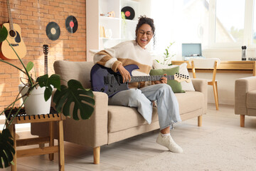 Happy young African-American woman playing electric guitar on sofa in living room