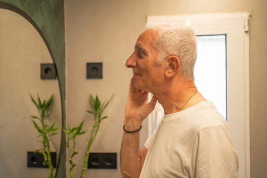 Side view of senior man examining his face reflection in a bathroom mirror, practicing self-care - Powered by Adobe