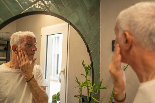 Senior man applying skincare product to his face, observing reflection in a bathroom mirror