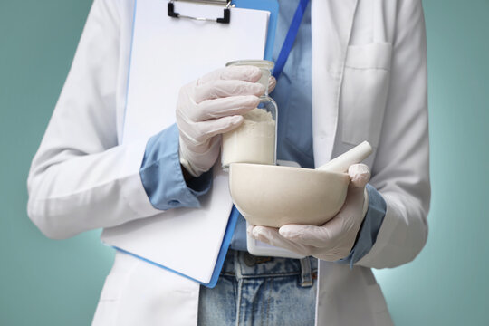 Young female scientist holding mortar with pestle, bottle and clipboard on turquoise background, closeup - Powered by Adobe