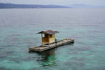 A simple, homemade bamboo raft with a small wooden shelter and a solar panel floating on clear, turquoise ocean water.