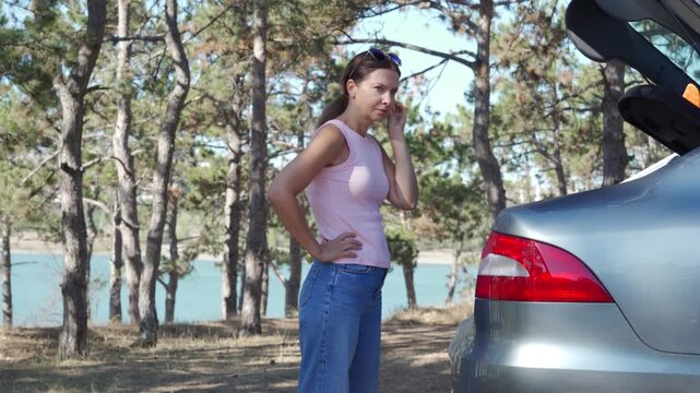 A woman and young boy stand near a silver car with an open trunk. They prepare for a family adventure in a sunny pine forest overlooking a lake.