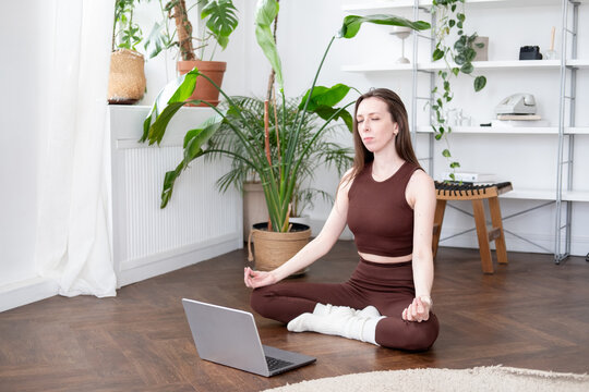  woman with long brown hair exercises with a laptop while wearing a sports bra and leggings, sitting on the floor against a backdrop of a white apartment and greenery. Exercises online via video call.