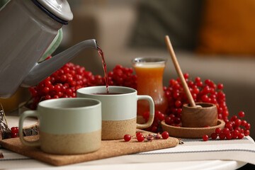 Pouring hot viburnum tea from teapot into cup on table in living room, closeup