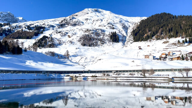 A majestic snowy mountain peak reflects perfectly in a calm lake, separated by a low-lying structure and alpine village houses - Powered by Adobe