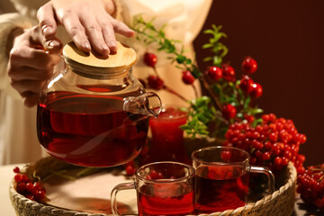 Young woman pouring hot viburnum tea from glass teapot into cup at home, closeup