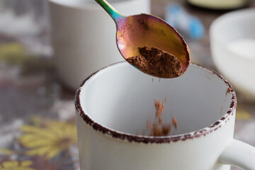 Preparing hot drink - putting cocoa powder into cup, closeup photo. Dropping cocoa powder into white mug from iridescent spoon, morning routine concept