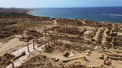 Aerial drone view over the ancient ruins of Sabratha, Libya, showing remains of temples, streets, and columns surrounded by desert vegetation and bordered by the deep blue Mediterranean waters
